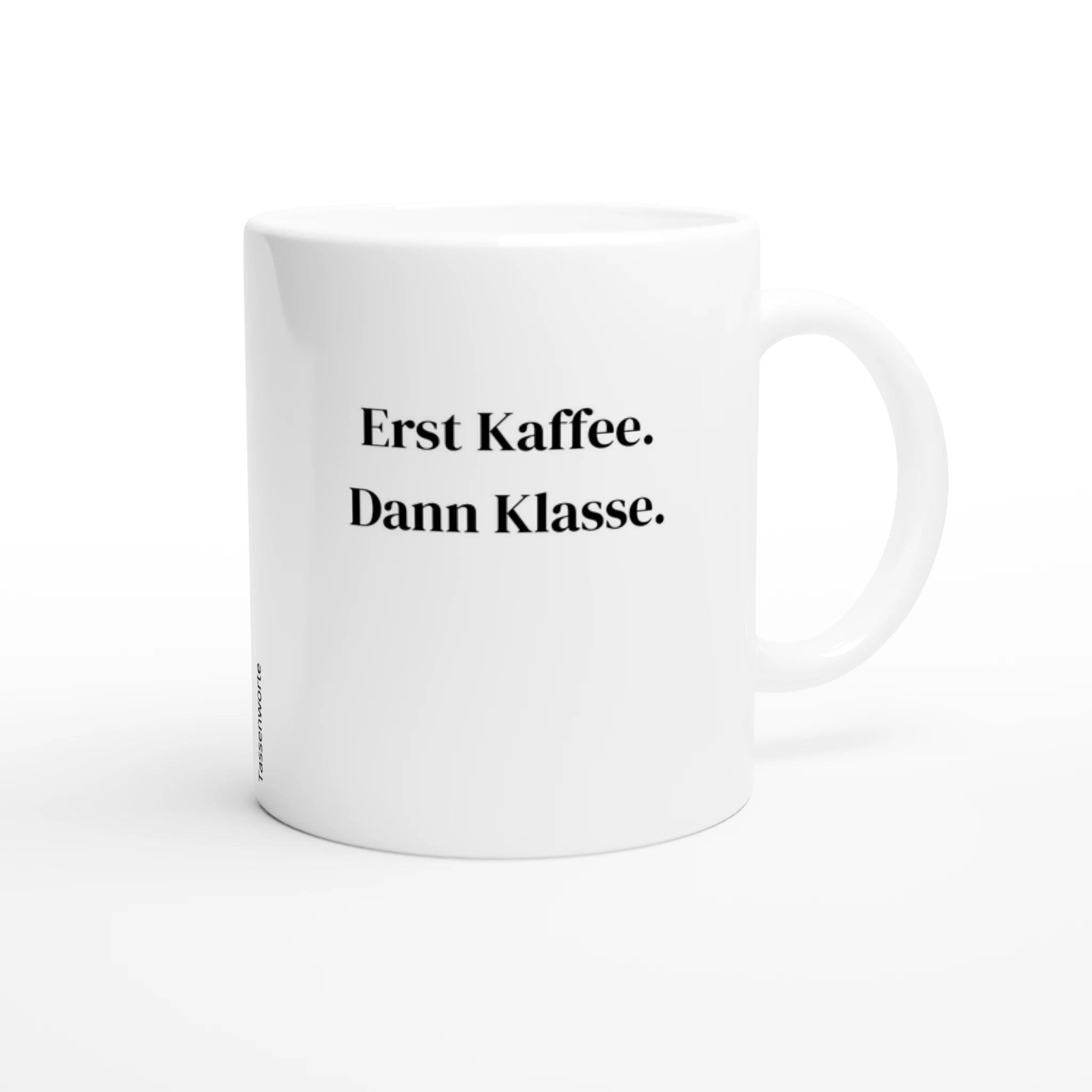 Eine schlichte, weiße Keramiktasse steht zentral auf einem neutralen, hellgrauen Untergrund. In der Mitte der Tasse ist in einer klassischen, schwarzen Serifenschrift der Text „Erst Kaffee. Dann Klasse.“ aufgedruckt. Die Tasse ist von oben schräg fotografiert, sodass die Öffnung und der Henkel auf der rechten Seite gut sichtbar sind.