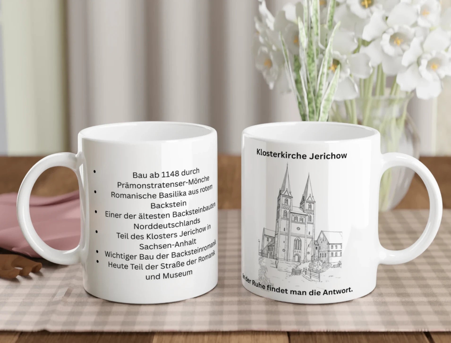 Zwei weiße Keramiktassen stehen auf einem gedeckten Holztisch vor einem Blumenstrauß. Die linke Tasse zeigt eine Auflistung historischer Fakten zur Klosterkirche Jerichow in schwarzer Schrift. Die rechte Tasse zeigt eine filigrane Strichzeichnung der Kirche mit zwei markanten Türmen, den Schriftzug „Klosterkirche Jerichow“ darüber und den Spruch „In der Ruhe findet man die Antwort“ am unteren Rand.