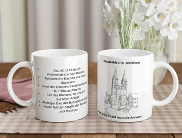 Zwei weiße Keramiktassen stehen auf einem gedeckten Holztisch vor einem Blumenstrauß. Die linke Tasse zeigt eine Auflistung historischer Fakten zur Klosterkirche Jerichow in schwarzer Schrift. Die rechte Tasse zeigt eine filigrane Strichzeichnung der Kirche mit zwei markanten Türmen, den Schriftzug „Klosterkirche Jerichow“ darüber und den Spruch „In der Ruhe findet man die Antwort“ am unteren Rand.