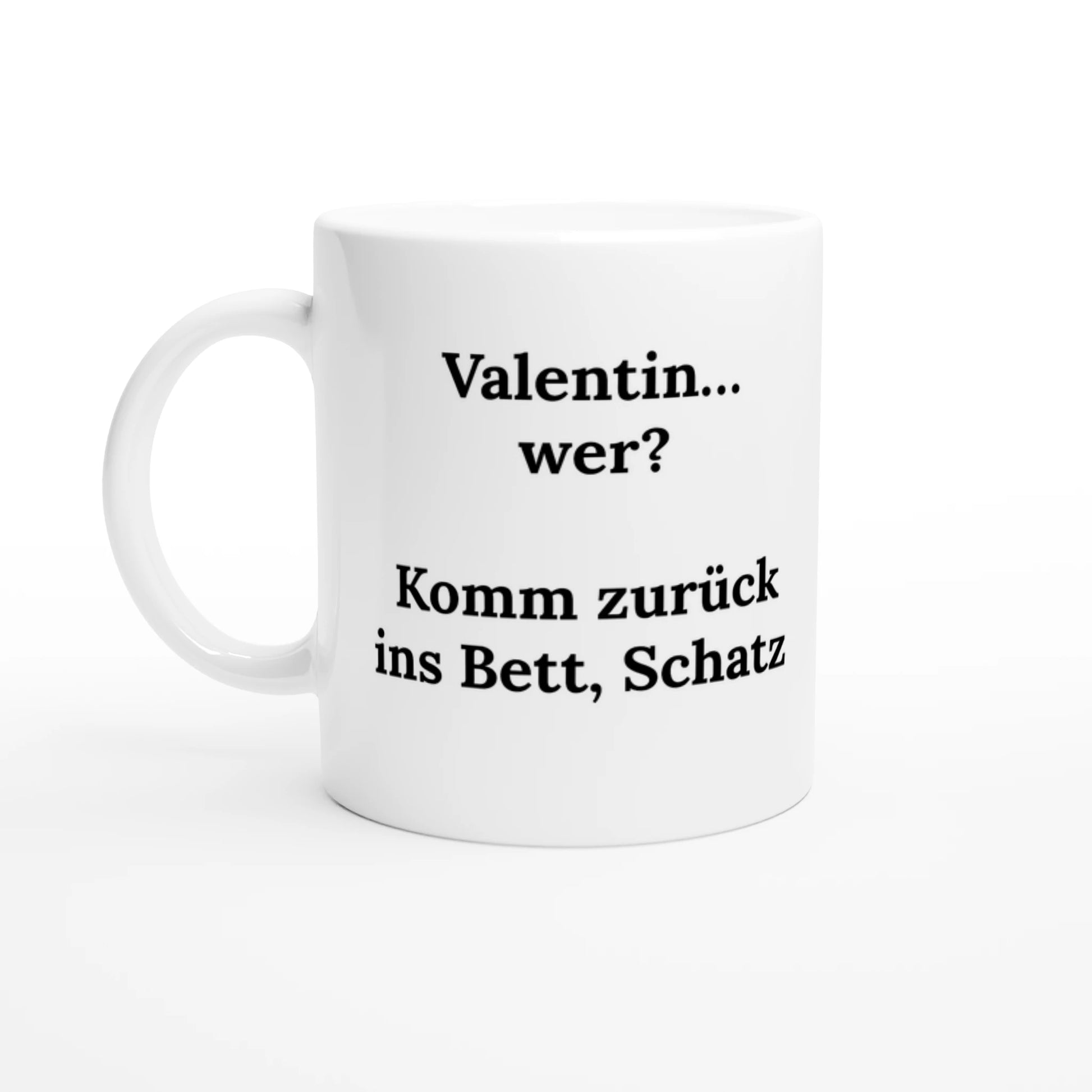 Eine weiße Kaffeetasse steht vor einem schlichten, hellen Hintergrund. Auf der Tasse ist ein schwarzer Text in einer Serifenschrift aufgedruckt. Der Text lautet: „Valentin... wer? Komm zurück ins Bett, Schatz“.