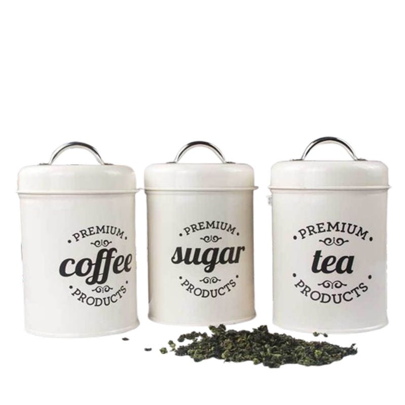 Drei cremefarbene Metall-Vorratsdosen mit silbernen Griffen und schwarzer Vintage-Beschriftung für "PREMIUM coffee PRODUCTS", "PREMIUM sugar PRODUCTS" und "PREMIUM tea PRODUCTS". Vor den Dosen liegt ein Haufen getrockneter Teeblätter. Von Tassenworte